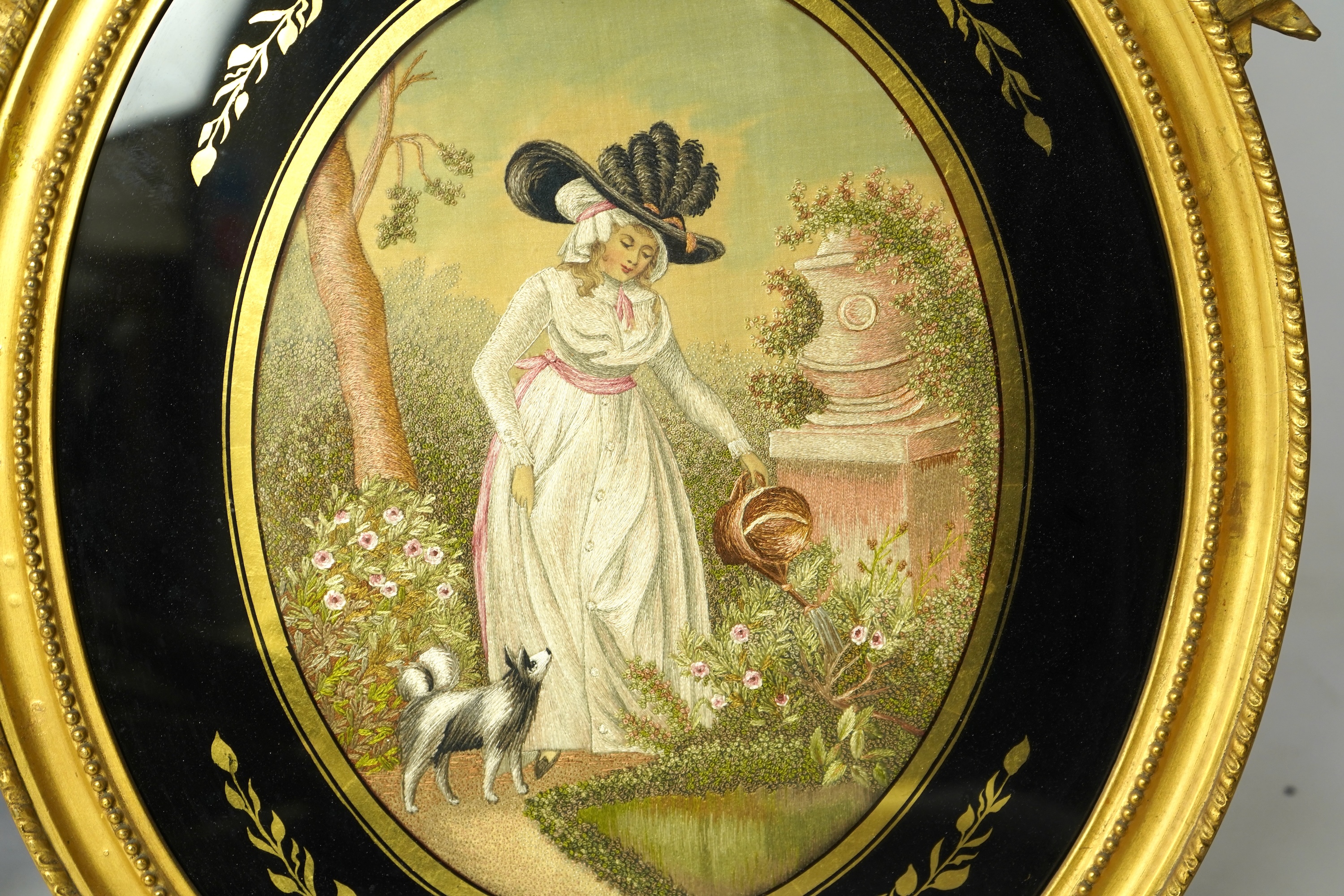 A pair of early 20th century oval silk embroideries, one of a mother and child dancing in a garden, the other of a lady in a large brimmed hat, watering flowers in by a classical stone urn, watched by her dog. Framed in